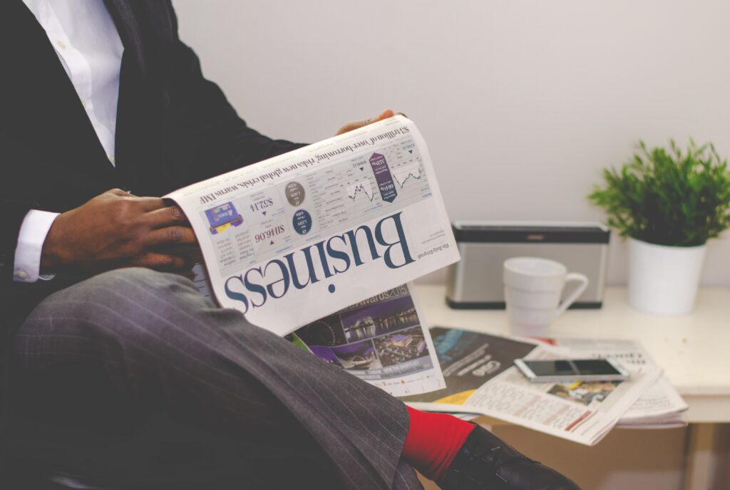 This image depicts a man sitting at a desk, focused on reading a business newspaper. He is dressed in business attire, with a suit jacket and a collared shirt. The newspaper is spread out in front of him, and he appears to be carefully reviewing an article. The man has a serious expression on his face, suggesting that he is taking the information in the newspaper seriously. The image conveys a sense of professionalism and dedication to staying informed about current business events.